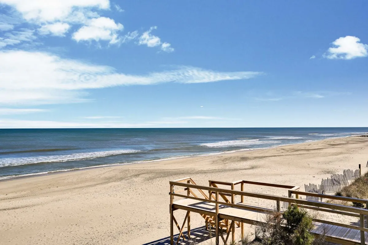 A broad view of Kure Beach with soft sand, gentle ocean waves, a winding boardwalk, and a wide-open sky. The image evokes classic beach activities such as building sandcastles, picnicking, and walking along the shore, with a family-friendly atmosphere.