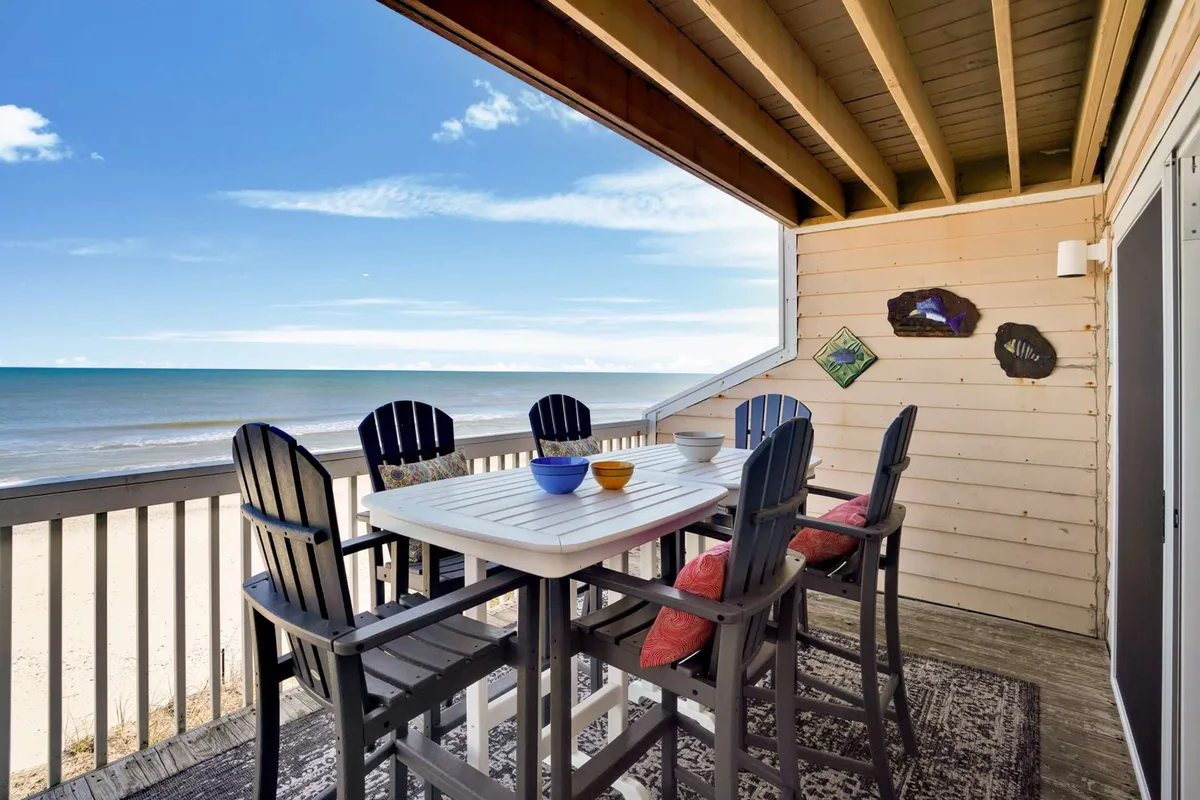An exterior scene highlighting a dining table set on a deck with colorful bowls, Adirondack chairs, and an expansive ocean view. The arrangement suggests a space perfect for outdoor meals, sunbathing, and social gatherings while enjoying the sunrise.