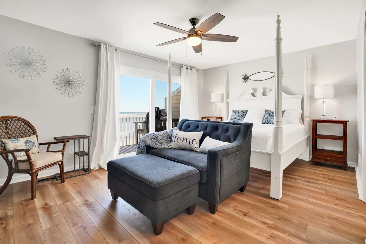 This image showcases a serene bedroom with a four-poster king bed, comfortable armchair, and plush ottoman. Sliding glass doors allow in abundant natural light and likely open to an oceanfront balcony, reinforcing the sense of a tranquil retreat. The cozy atmosphere is complemented by neutral, calming décor and inviting textures.