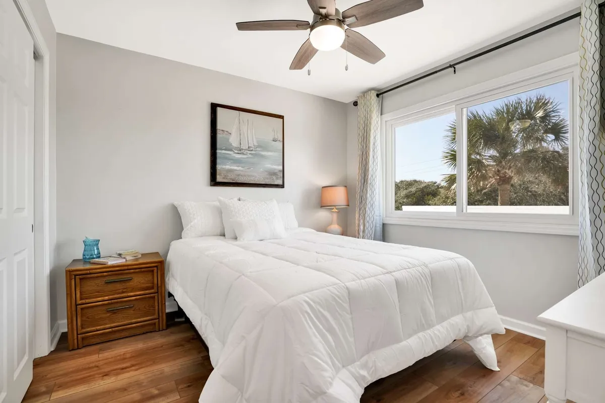 A queen bedroom with crisp white bedding, a wooden nightstand, and beach-themed artwork. A window frames a view of swaying palm trees, bringing the outdoors in. The room exudes a feeling of quiet comfort and is bathed in soft, natural light.