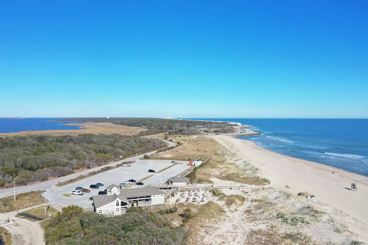 A lively exterior view featuring a beach, ocean, parking area, a restaurant, and a walkway. The scene captures people enjoying the beach, dining, and exploring nearby trails in a relaxed, inviting atmosphere.