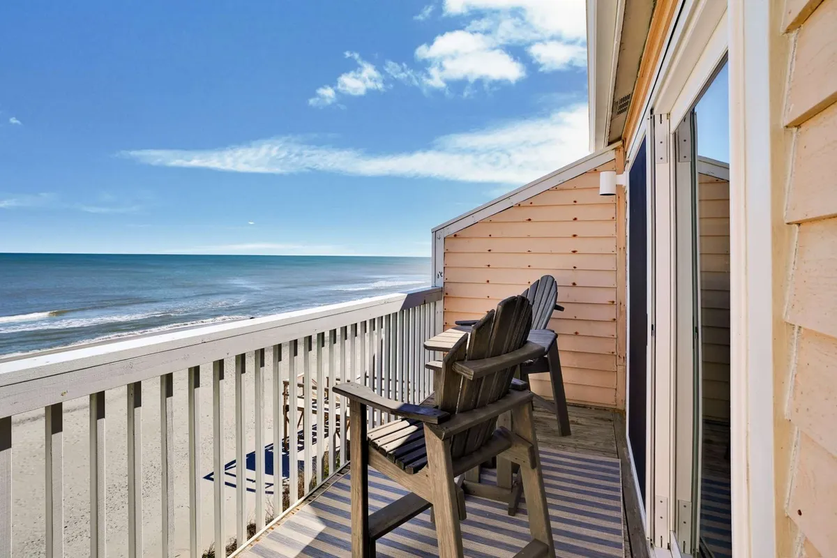 The image shows a serene outdoor scene at an oceanfront condo in Kure Beach, North Carolina. Wooden chairs are placed on a balcony or deck, facing an expansive view of the sparkling ocean under a clear blue sky. The atmosphere is peaceful, inviting guests to relax, sip coffee, read a book, or watch the sunrise and sunset as the ocean breeze drifts by.