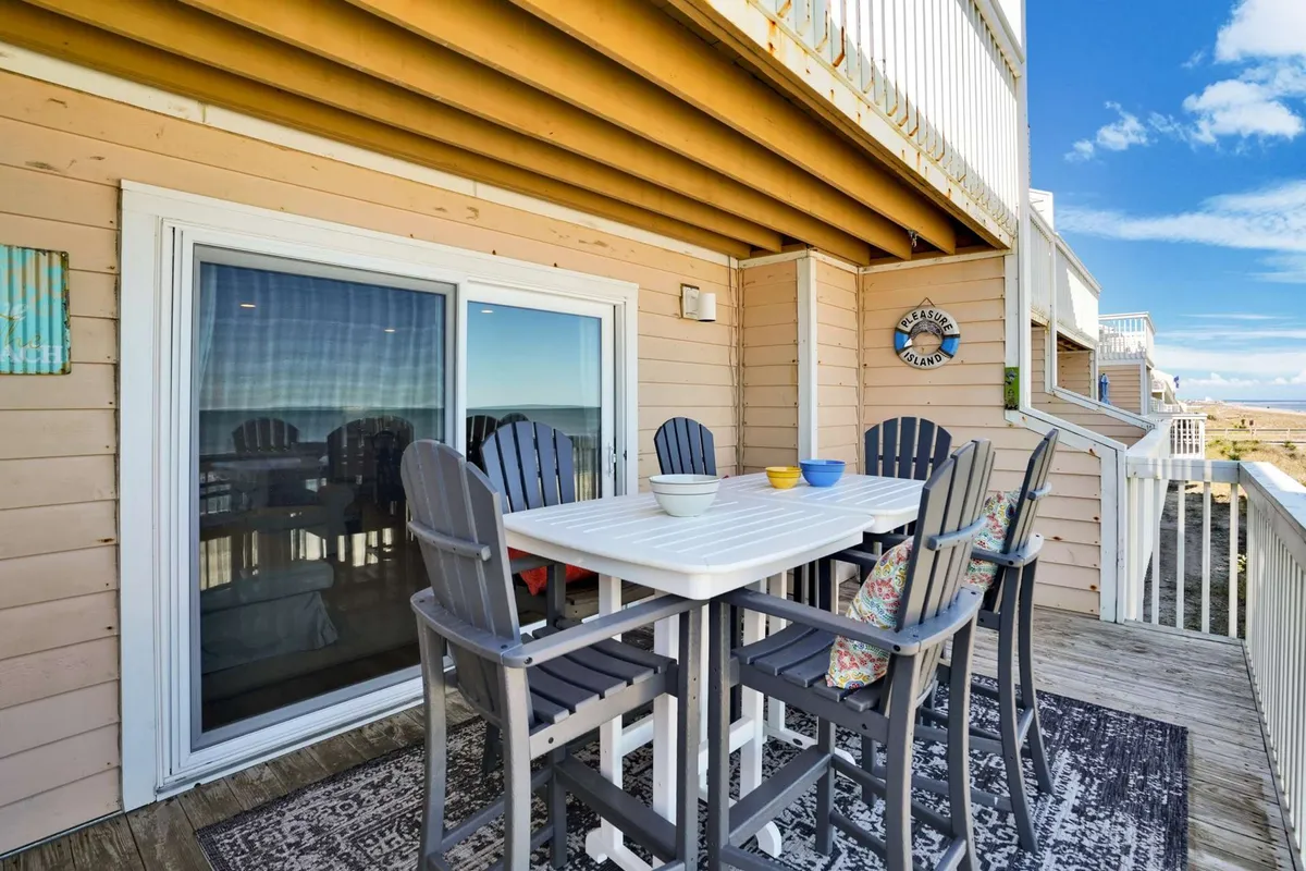 An inviting outdoor dining table with Adirondack chairs faces a sweeping beach view. The table is set up for al fresco dining, and the setting exudes casual comfort, perfect for meals or game nights as a group.