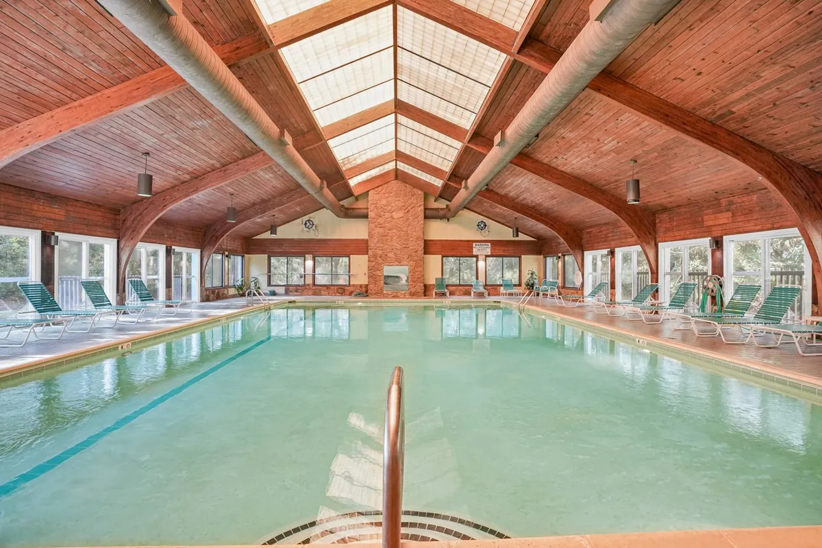 A bright indoor swimming pool with wooden beams, large windows, and comfortable lounge chairs. The space feels tranquil and rejuvenating, with sunlight filtering in to create a warm, inviting environment for relaxation and year-round swimming.