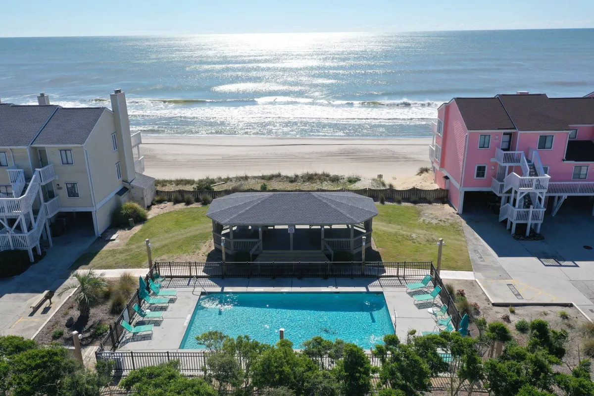 This image captures a scenic view of the resort's swimming pool with a gazebo, set just in front of the beach and ocean, and surrounded by the resort community buildings. The pool area is bathed in natural light and features inviting spaces for swimming, sunbathing, and gathering as a family.
